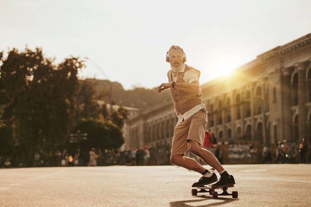 Portrait of handsome man in white shirt on sunset evening in summer. Stylish guy ride on skateboard on city street. Urban male lifestyle on buildings background - Photo, Image