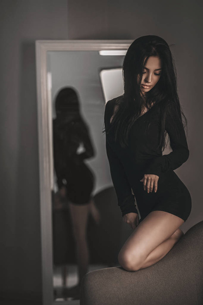 Seductive woman portrait of trendy woman in black dress on armchair. Stylish fashion girl model posing near mirror in bedroom - Foto, Imagen