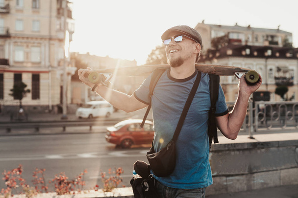 Boldog ember napszemüvegben, kék ingben és sapkában gördeszkával az út mellett, utcai háttérben. Egy kültéri portré egy stílusos fickóról. Városi életmód - Fotó, kép