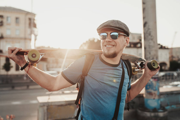 Skater pozowanie o zachodzie słońca na ulicy miasta. Stylowy hipster człowiek z longboard spaceru w pobliżu drogi na tle budynków. Portret brody sport męski model w stylu miejskim - Zdjęcie, obraz