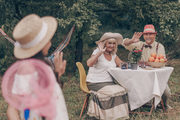 幸せな祖母と祖父はテーブルに座ってレモネードを飲み、庭の孫娘に手を振っています。蝶のネットを持っている美しい若い孫娘。彼女はサンドレスと帽子をかぶっている。. - 写真・画像