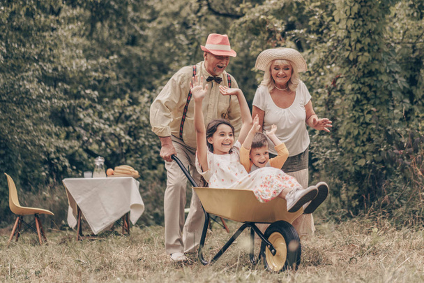 Бабусі і дідусі в парку з онуками граються з візком. Родина розважається в саду. Старі люди раді бачити онука та онука
 - Фото, зображення