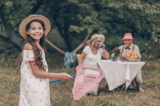 glückliches junges Mädchen, das mit einem Netz in einer Sonnenhose und einem Hut im Garten posiert. Porträt einer lächelnden Enkelin. alte Großeltern beobachten und genießen den Häuptling - Foto, Bild
