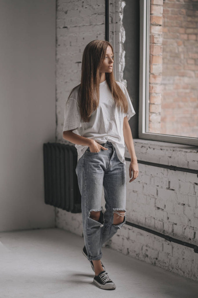 Seductive portrait of young sexy girl with brunette hair in shirt, jeans and sneakers on white background in room near window - Foto, immagini