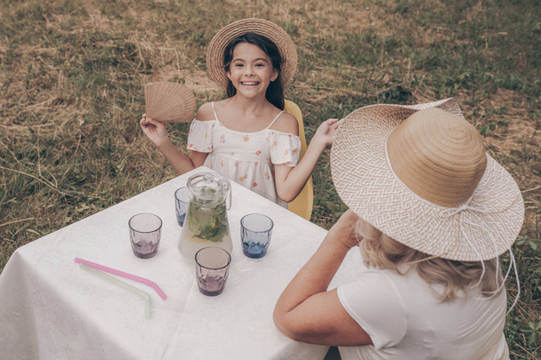 Yelpazesi ve şapkası olan küçük bir kız yaşlı bir kadınla masaya oturur ve limonata içer. Mutlu bir aile. Güzel torun, doğada bahçede oturmak için büyükannesini ziyarete geldi. Ebeveynlerin ve çocukların oranı - Fotoğraf, Görsel