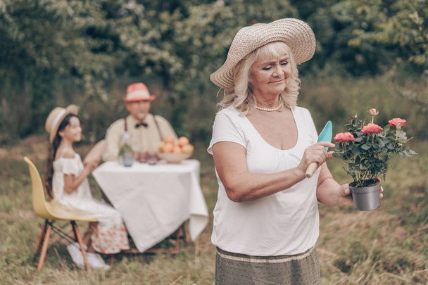 La abuela feliz hace una sorpresa para su nieta. El cuidado del medio ambiente, quiere plantar una flor. En el fondo se sienta una niña y un abuelo en una mesa, sonriendo y bebiendo fruta de limón. Una familia feliz
 - Foto, imagen