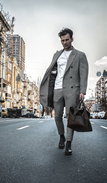 Divatmodell férfi sétál az úton közel az üzleti központhoz. Utcai fotózás. Meleg szürke kabátban, fehér pulóverben, fekete cipőben, barna bőrtáskában.. - Fotó, kép