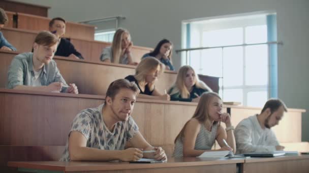 Moderne studenten, mannen en vrouwen, zitten op bureaus in een groot klaslokaal en schrijven een lezing notities. - Video