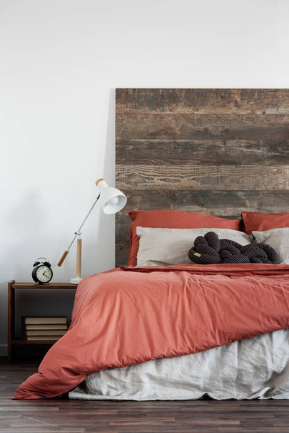 Trendy white lamp next to black clock on wooden nightstand next to king size bed with wooden headboard - 写真・画像