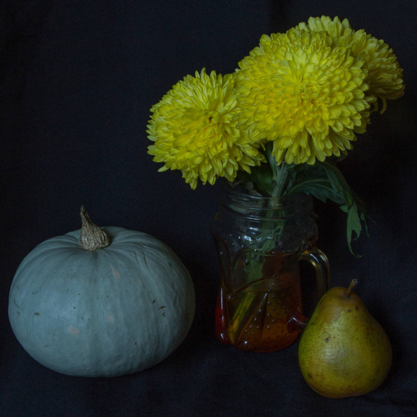 Yellow chrysanthemums in a glass mug, green pumpkin, pear on a black background, autumn still life - 写真・画像