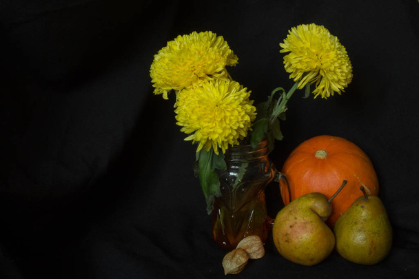 Keltaiset krysanteemit lasimukissa, oranssi kurpitsa, kaksi päärynää ja kaksi physalista mustalla taustalla, syksyn asetelma
 - Valokuva, kuva