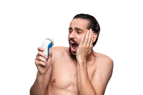 Asustado hombre barbudo con bigote mira a la maquinilla de afeitar eléctrica de pie desnudo aislado sobre fondo blanco. Concepto de tratamiento matutino y afeitado. Hora de cortarte la barba. Rutina matinal
. - Foto, Imagen