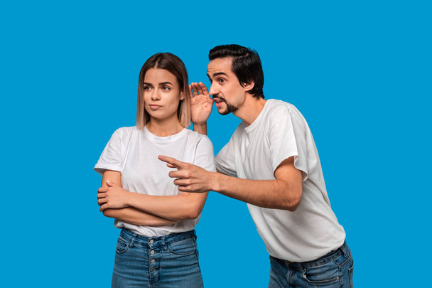 Bearded man with mustaches in white t-shirt tells secret to a young blond girl in white t-shirts and blue jeans standing isolated over blue background. Woman looks surprised. Secret telling. - Photo, Image