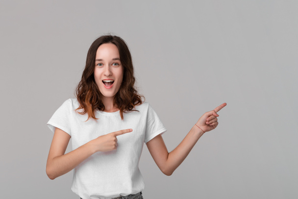 Pretty brunette woman in a white tee pointing at the empty space in a right side of a frame standing isolated over grey background. Copy space of advertising. - Фото, изображение