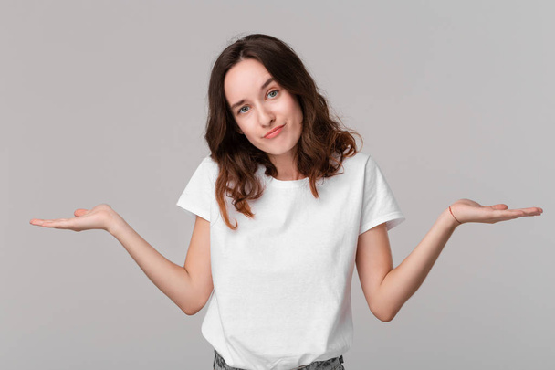 Pretty brunette woman in a white tee shrugging her shoulders beacause she is indignant  standing isolated over grey background. Copy space of advertising. - Fotografie, Obrázek