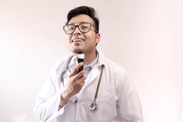 male indian doctor in white coat and stethoscope trimming beard with trimmer. doctor styling theme image - Photo, Image