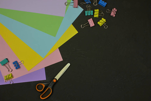 Top view of multicolor paper, paper clips and scissors on dark background with copy space. Back to school, education concept, school accessories. - Photo, Image