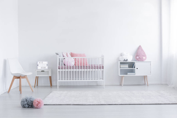 Pink pillow on cabinet and white chair in simple kid's room interior with carpet and cradle. Real photo - Foto, afbeelding