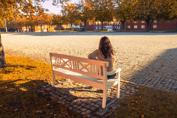 Beautiful lonely girl sitting on a white bench in the autumn - Photo, Image