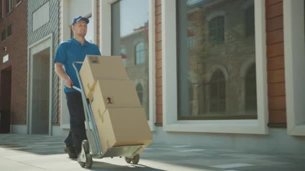 Happy Young Delivery Man Pushes Hand Truck Trolley Full of Cardboard Boxes and Packages For Delivery. Professional Courier Working Efficiently and Quickly. In the Background Stylish Modern Urban Area - Materiał filmowy, wideo