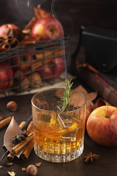 Un verre de whisky alcoolisé avec une branche de romarin avec de la fumée et des pommes, des bâtonnets de cannelle. Concept d'automne magique atmosphère détendue et confortable
. - Photo, image