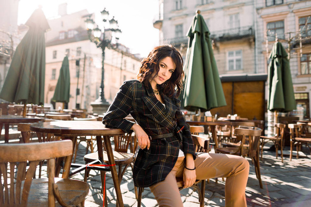 Beautiful happy smiling girl with dark hair, nude makeup, wearing stylish black jacket posing in street cafe. Outdoor portrait, day light. Female autumn fashion concept. Copy, empty space for text - 写真・画像