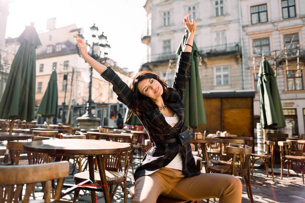 beautiful woman sitting in a cafe on a city street, fashion style, hands up, wake up in the morning, enjoy sunny day - 写真・画像