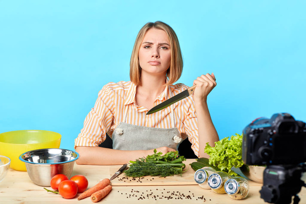 Attractive lifestyle blogger sits with big knife, has thoughtful expression - Foto, imagen