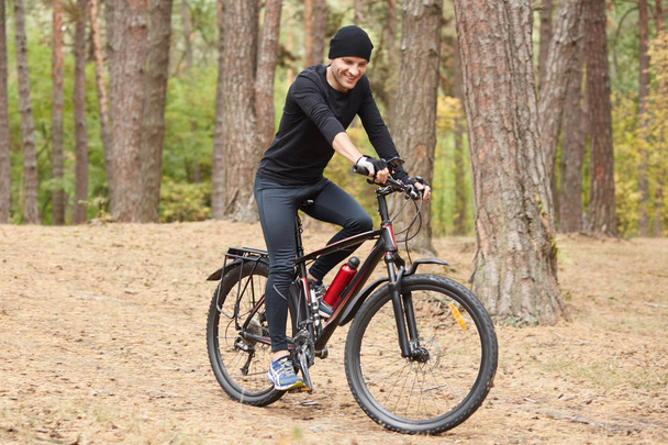 Pictiure of biker riding through forest, wearing black cycling clothes, cap and sneakers, having happy facial expression, sporty male spending leisure time in open air, riding around trrees. - Fotoğraf, Görsel