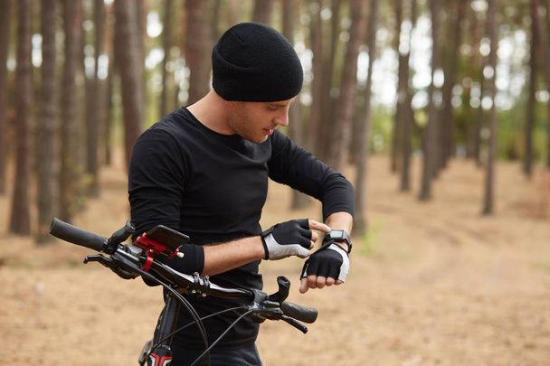 Outdoor shot of handsome man using his smart watch, riding bike in forest, wearing black sportwear and cap, looks athis device, spending leisure time inopen air, enjoys fresh air and beautiful nature. - Foto, imagen