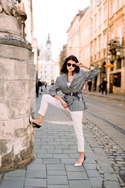 young brunette woman in elegant business clothes and stylish sunglasses, sexy posing near old stone wall of the building, outdoor shot - Zdjęcie, obraz