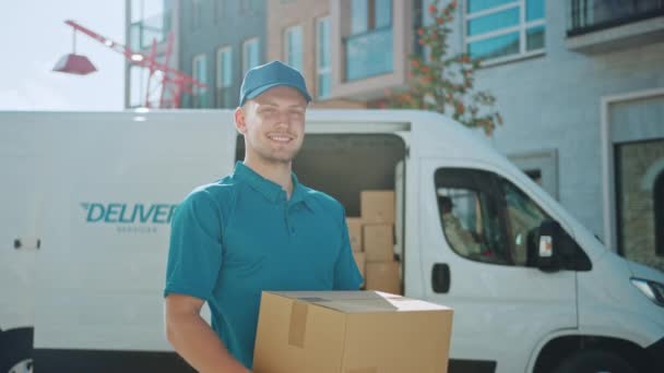 Portrait of Handsome Delivery Man Holds Cardboard Box Package Standing in Modern Stylish Business District with Delivery Van in Background. Smiling Courier On Way to Deliver Postal Parcel to Client - Imágenes, Vídeo