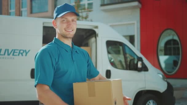 Portrait of Handsome Delivery Man Holds Cardboard Box Package Standing in Modern Stylish Business District with Delivery Van in Background. Smiling Courier On Way to Deliver Postal Parcel to Client - Felvétel, videó