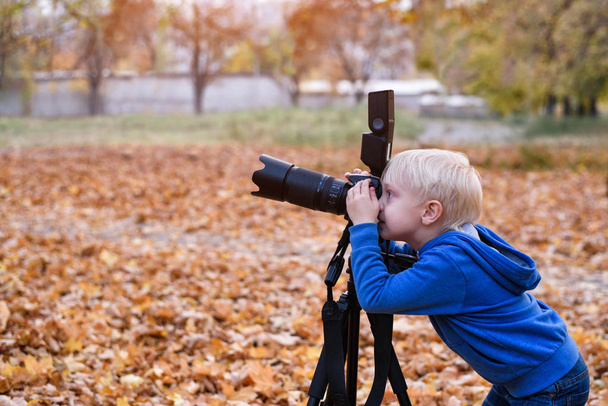 Pieni vaalea poika ampuu suurella SLR-kameralla jalustalla. Valokuvaussessio syksyn puistossa
 - Valokuva, kuva