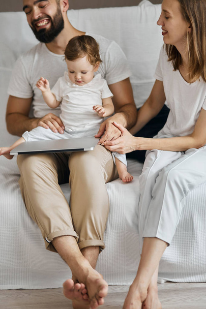 Young parents with laptop computer on knees playing with baby - 写真・画像