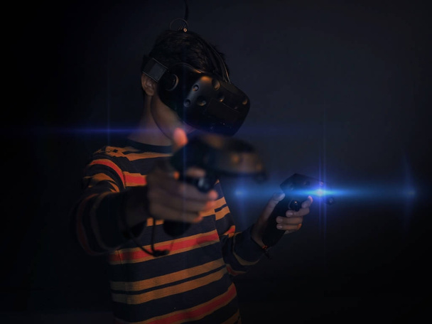 Νεαρό ασιατικό αγόρι κρατώντας και φορώντας VR παιχνίδι σετ στο μαύρο φόντο. Επιλεγμένη εστίαση. - Φωτογραφία, εικόνα