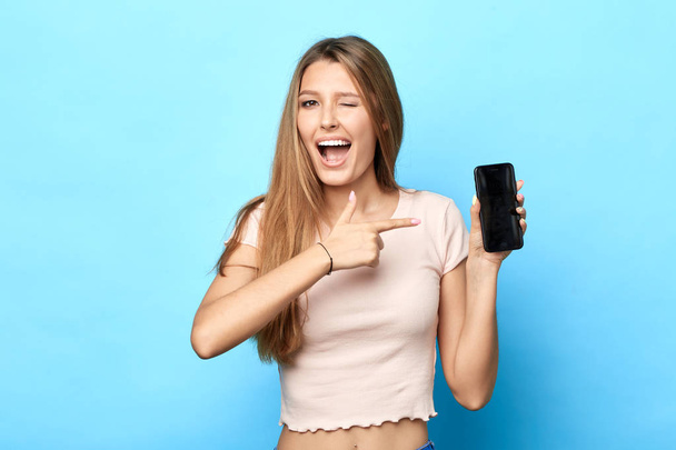 funny crazy attractive girl is pointing on smartphone with finger standing on blue background. - Fotó, kép