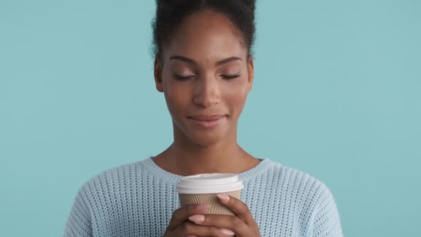 Όμορφη Αφροαμερικανή κοπέλα σε άνετο πουλόβερ ονειρικά μυρίζει καφέ και ευτυχώς κοιτάζοντας στην κάμερα πάνω από το μπλε φόντο - Πλάνα, βίντεο