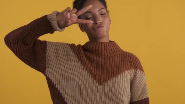 Menina americana muito africana em camisola de malha brincando mostrando gesto de paz sobre fundo amarelo
 - Filmagem, Vídeo