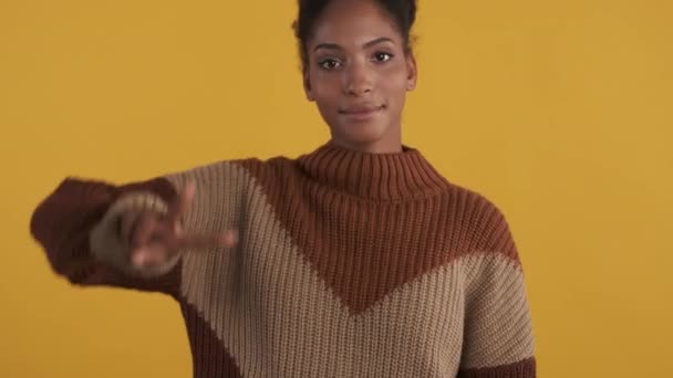 Портрет веселої афро-американської дівчини в в'язаному светрі з радістю показує V-знак на камері на жовтому фоні - Кадри, відео
