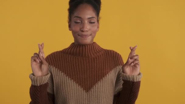 Portret podekscytowanej afrykańskiej dziewczyny z skrzyżowanymi palcami, miejmy nadzieję modlącej się przed kamerą na żółtym tle - Materiał filmowy, wideo
