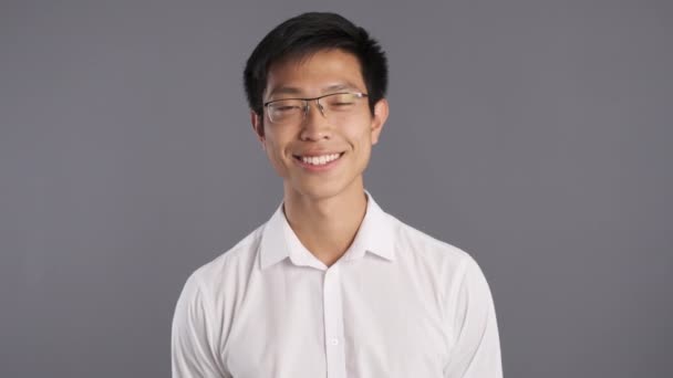 Retrato de joven sonriente hombre asiático en gafas felizmente mirando en cámara sobre fondo gris
  - Metraje, vídeo