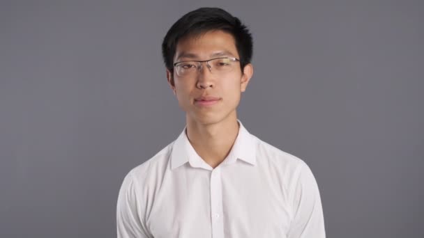 Portrait of young cheerful asian man in eyeglasses happily looking in camera over gray background  - Footage, Video