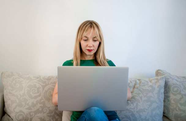 European woman blonde in glasses and a green T-shirt works at home on a laptop. A woman freelancer feels joy and satisfaction from her work - Photo, Image