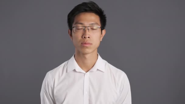 junger ernster asiatischer Mann mit Brille zeigt selbstbewusst Schweigegeste vor der Kamera vor grauem Hintergrund  - Filmmaterial, Video