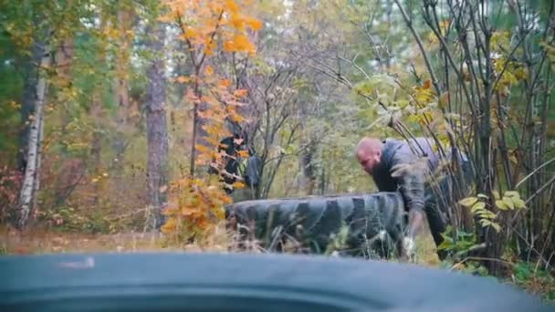 Wytatuowany kulturysta w rękawiczkach popycha opony ciężarówki na trening naziemny w jesiennym lesie - Materiał filmowy, wideo