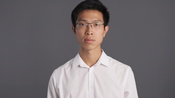 Young serious asian man in eyeglasses pointing fingers on himself and showing no gesture on camera over gray background  - Footage, Video
