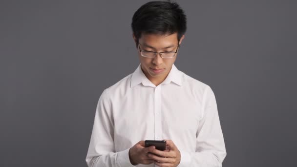 Jovem sorrindo asiático homem em óculos alegremente usando celular sobre fundo cinza
  - Filmagem, Vídeo