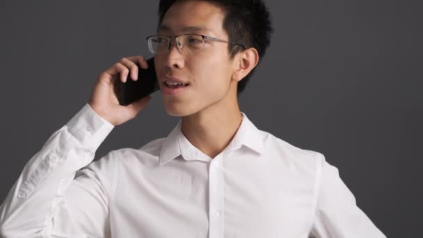 Jovem atraente positivo asiático homem em óculos alegremente falando no celular sobre fundo cinza
  - Filmagem, Vídeo