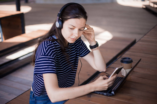 A girl student with a smartphone, sits at a laptop and listens carefully in headphones, learns foreign languages. - Photo, Image